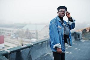 hombre afroamericano con chaqueta de jeans, boina y anteojos posados en un techo abandonado. foto