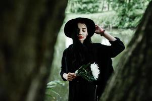 Sensual girl all in black, red lips and hat. Goth dramatic woman hold white chrysanthemum flower at hand against silent lake. photo