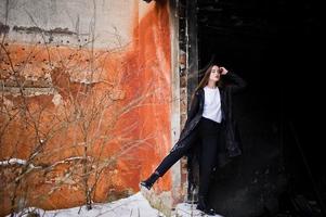 Fashionable long legs brunette model in long black cloak posed outdoor at winter day against old grunge wall. photo