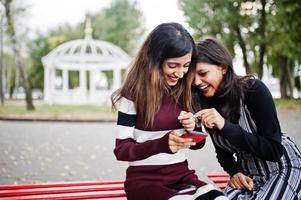 Portrait of two young beautiful indian or south asian teenage girls in dress sitting on bench and use mobile phone. photo