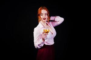 Gorgeous red haired girl in pink blouse and red skirt with glass of wine at hand isolated on black. photo