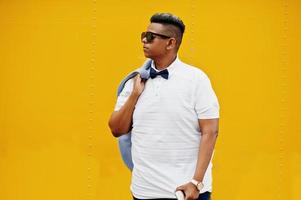 Close up portrait of stylish arabian man at sunglasses and bow tie posed against yellow background with mobile phone. Arab model boy. photo