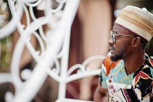 Handsome afro american man wearing traditional clothes, cap and eyeglasses sitting at white carriage. photo