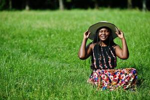Portrait of gorgeous african american woman 20s in summer hat sitting at green grass in park. photo