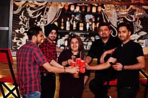 Group of indian friends having fun and rest at night club, drinking cocktails, clinking and cheers near bar counter. photo