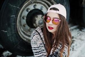 Brunette stylish casual girl in cap and sunglasses sitting against truck wheels. photo