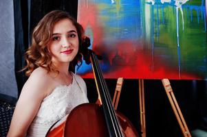 Pretty young gilrl musician in white dress with double bass next to her art painting. photo