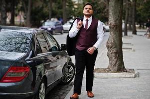 hombre de negocios indio con estilo en ropa formal de pie contra el coche de negocios negro en la calle de la ciudad. foto