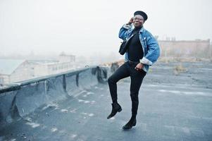 hombre afroamericano con chaqueta de jeans, boina y anteojos posados en un techo abandonado. foto