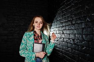 Stylish blonde girl in jacket and jeans with notebook diary at hands against brick black wall at studio. photo