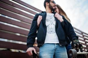 Cool multiracial couple hugs together with longboard against wooden fence. photo