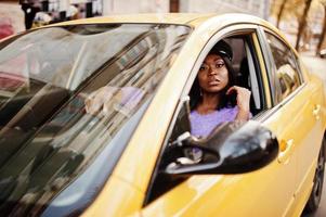 mujer afroamericana con vestido violeta y gorra posada en un auto amarillo. foto