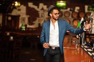 Handsome well-dressed arabian man with glass of whiskey and cigar posed at pub. photo