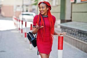 Cute and slim african american girl in red dress with dreadlocks and backpack posed outdoor and looking at mobile phone on street. Stylish black model. photo