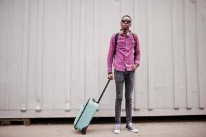 African american man in checkered shirt, sunglasses and earphones with suitcase and backpack. Black man traveler against wall. photo