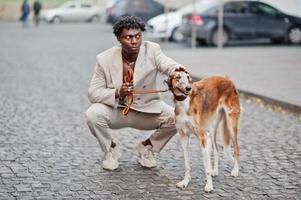 Stylish afro man in beige old school suit with Russian Borzoi dog. Fashionable young African male in casual jacket on bare torso. photo