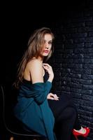 Handsome brunette girl wear on black and green jacket in red high heels, sitting and posing on chair at studio against dark brick wall. Studio model portrait. photo