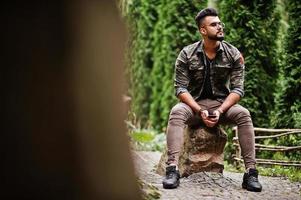 Awesome beautiful tall ararbian beard macho man in glasses and military jacket posed outdoor, sitting on stone. photo