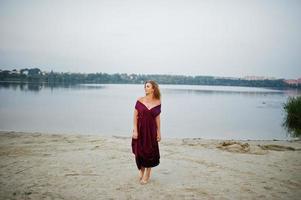 Blonde sensual barefoot woman in red marsala dress posing against lake on sand. photo