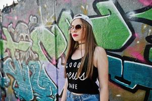 Stylish casual hipster girl in cap, sunglasses and jeans wear against large graffiti wall. photo