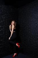 Handsome brunette girl wear on black and red high heels, posing at studio against dark brick wall. Studio model portrait. photo