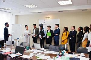 Mixed race business coach presenting report standing near whiteboard pointing on sales statistic shown on diagram and chart teach diverse company members gathered together in conference room. photo