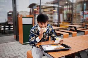 Attractive and handsome stylish indian man wear on shirt eat burger and french fries in the terrace of modern lounge cafe at his work break. photo