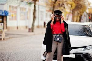 African american fashion girl in coat, newsboy cap and sunglasses posed at street against white business car. photo