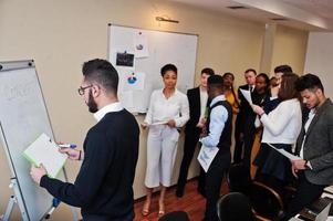 Mixed race business coach presenting report standing near whiteboard pointing on sales statistic shown on diagram and chart teach diverse company members gathered together in conference room. photo