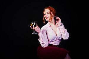 Hermosa chica pelirroja con blusa rosa y falda roja con una copa de vino a mano aislada en negro. foto