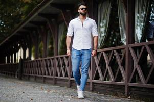 Stylish tall arabian man model in white shirt, jeans and sunglasses walking at street of city. Beard attractive arab guy. photo