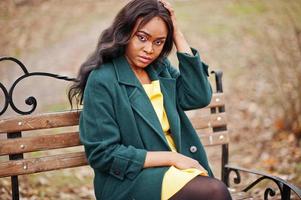 elegante mujer afroamericana con abrigo verde y vestido amarillo sentada en un banco. foto