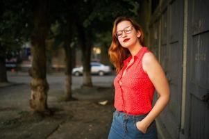 Attractive redhaired woman in eyeglasses, wear on red blouse and jeans skirt posing. photo