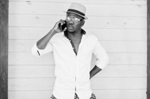 Stylish african american man in white shirt and colored pants with hat and glasses posed outdoor speaking mobile phone. Black fashionable model boy. photo