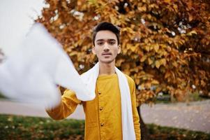 Indian stylish man in yellow traditional clothes with white scarf posed outdoor against autumn leaves. photo