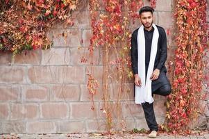 Indian stylish man in black traditional clothes with white scarf posed outdoor against red leaves wall. photo