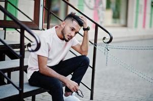 Stylish indian beard man at pink t-shirt. India model sitting on stairs outdoor at streets of city. photo