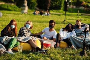 joven grupo multiétnico de personas viendo películas en poof en el cine al aire libre usando una máscara durante la cuarentena del coronavirus covid. foto