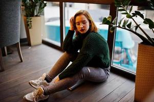 Cheerful young beautiful redhaired woman in glasses, green warm wool sweater, using her notebook, while sitting on cafe against window. She sit on floor and think about something. photo