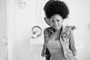 Attractive african american woman with afro hair wear on skirt and jeans jacket, posed at white room. Fashionable black model. photo