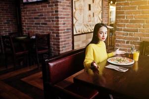 Funny brunette girl in yellow sweater eating pizza at restaurant. photo