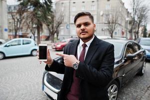 Stylish indian businessman in formal wear standing against black business car on street of city and show finger to mobile phone screen. photo