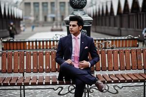 elegante modelo de hombre macho indio con traje y corbata rosa posado en el día de invierno y sentado en un banco. foto