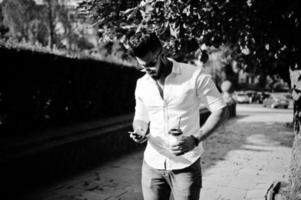 Stylish tall arabian man model in white shirt, jeans and sunglasses walking at street of city. Beard attractive arab guy with cup of coffee and mobile phone at hands. photo