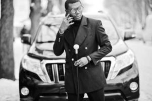 Stylish african american gentleman in elegant black jacket, holding retro walking stick as cane flask or tippling cane. Rich fashionable afro man against business suv car speaking on mobile phone. photo