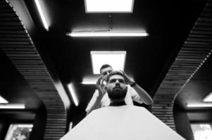 Young bearded man getting haircut by hairdresser while sitting in chair at barbershop. Barber soul. photo