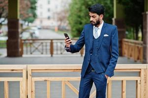 hombre de negocios indio de barba elegante con bindi en la frente, vestido con traje azul posado al aire libre y haciendo selfie en el teléfono móvil. foto