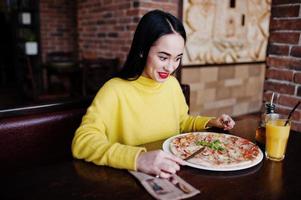 chica morena divertida en suéter amarillo comiendo pizza en el restaurante. foto