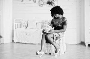 Afican american women in brilliant glitter sequins turqoise dress sitting on chair at white room with decorative rose at hands. photo