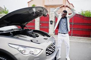 Stylish and rich african american man stand in front of a broken suv car needs assistance looking under opened hood. photo